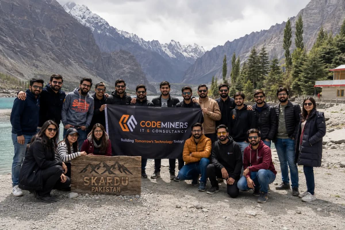 Entire CodeMiners engineering team group photo at Skardu, Pakistan — holding CodeMiners banner with Karakoram mountain range and glacial lake in background, annual team retreat